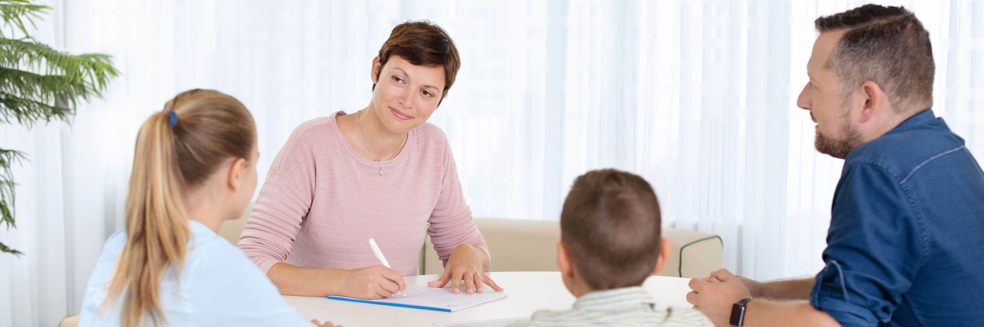 Eine Psychiaterin sitzt mit zwei Kindern und deren Vater am Tisch und macht sich Notizen